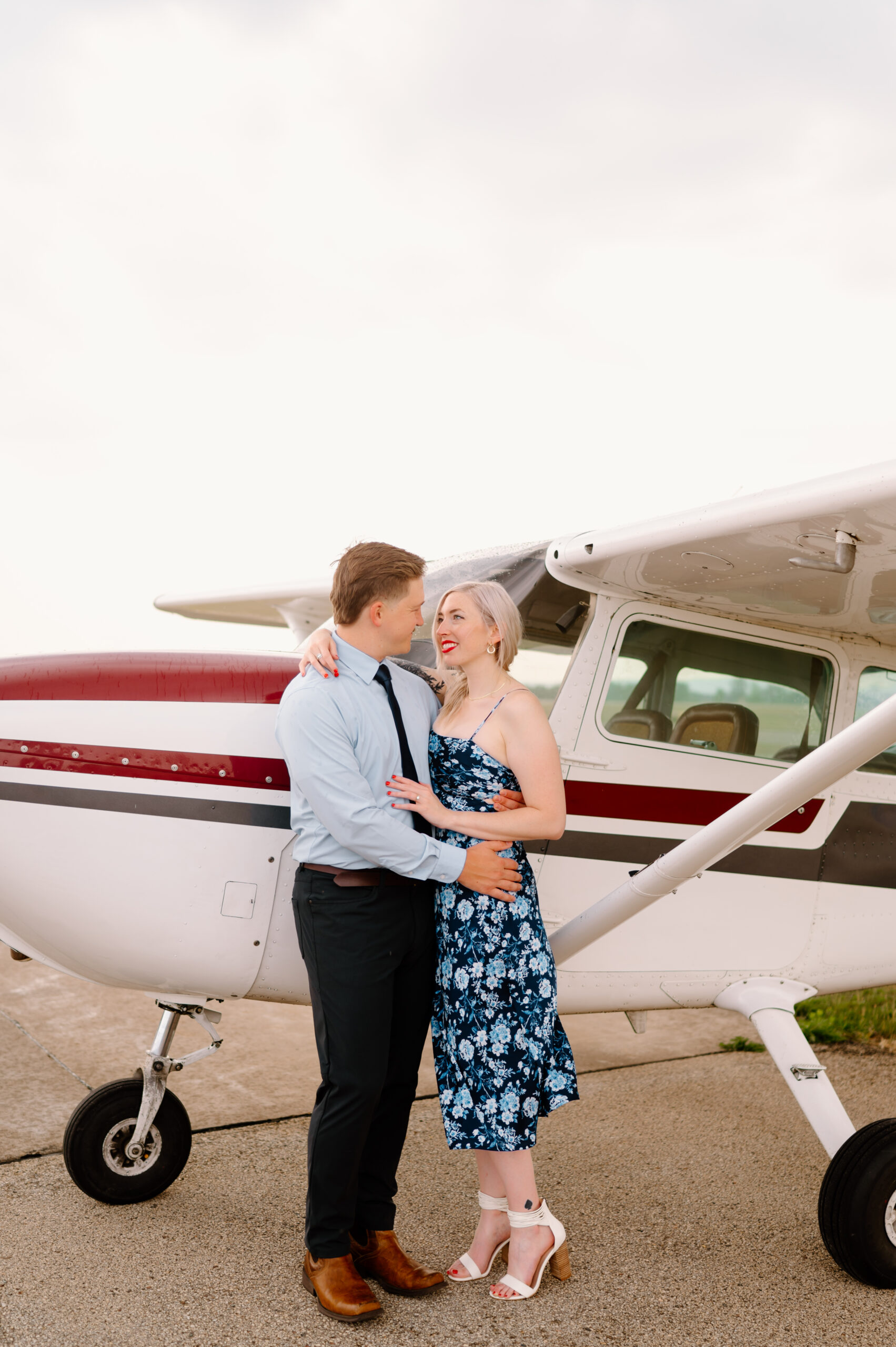 Unique engagement session of a couple with their arms wrapped around each other standing in front of their single engine plane.