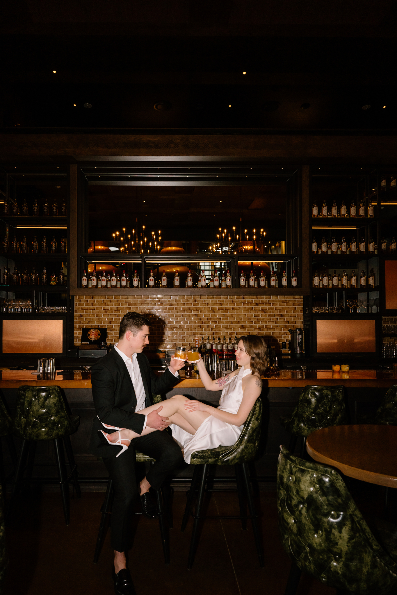 Couple sitting at a nice bar. He is wearing a black suit and she is wearing a long white dress.