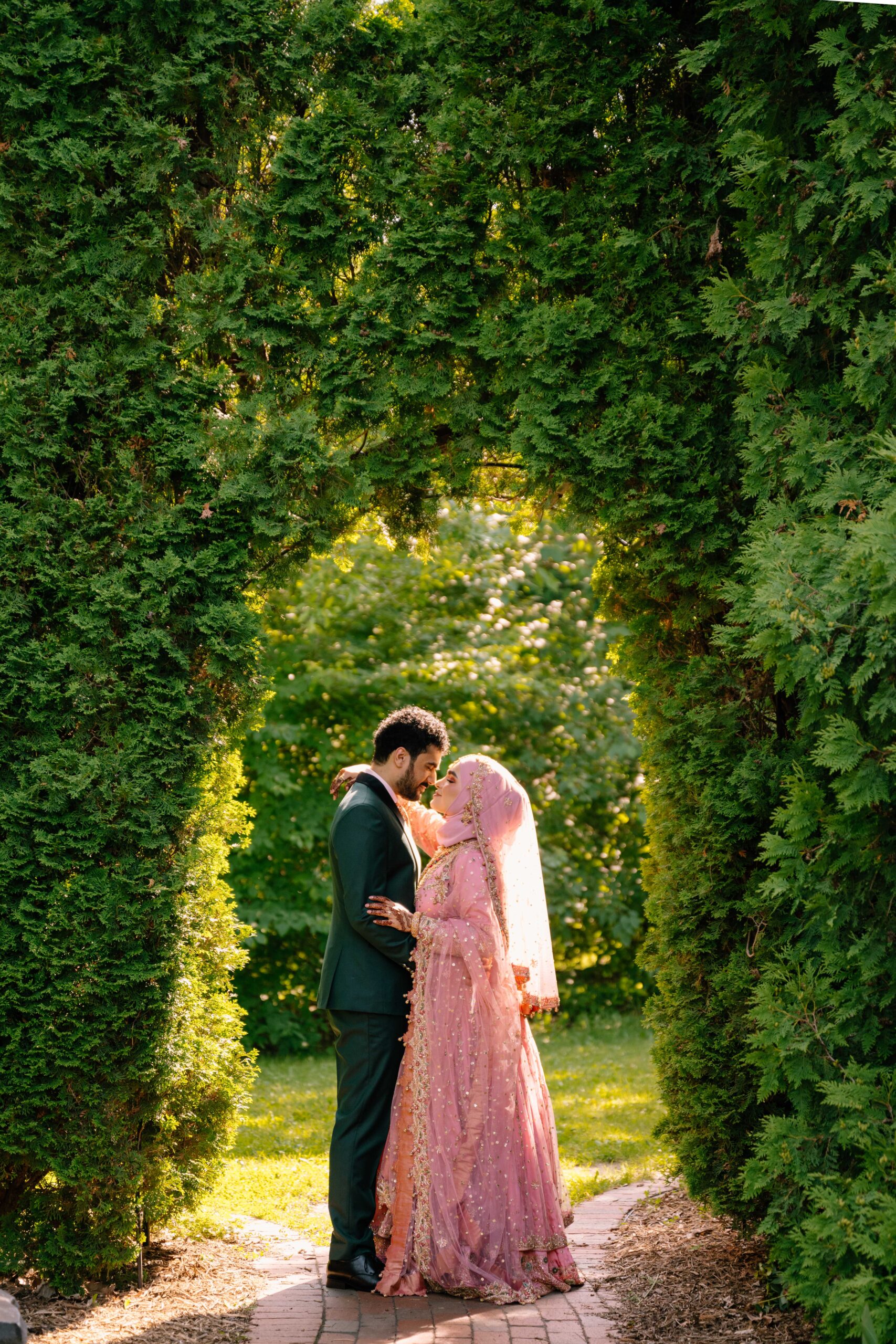 Bride and Groom embracing under an arch filled with greenery.